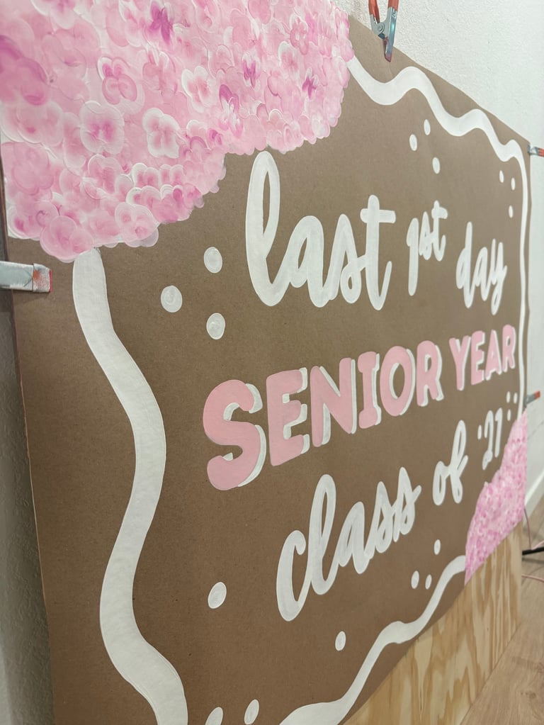 Brown cardboard cutout sign with pink pom-poms reading Last First Day Senior Year Class of 2027