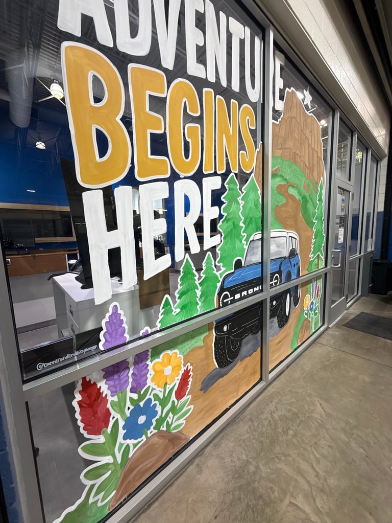 Storefront window display with yellow and white Adventure Begins Here text, featuring illustrated camping van, pine trees, and colorful flowers