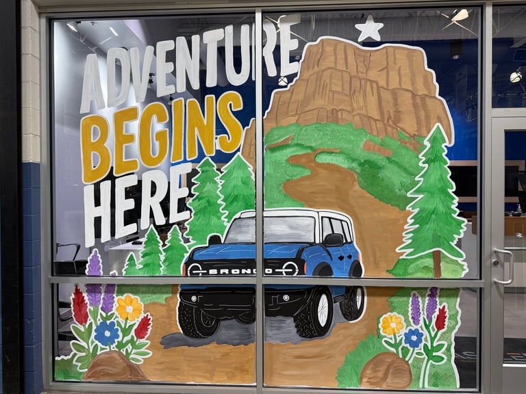 Window mural with Adventure Begins Here text, illustrated 4x4 truck, desert landscape, mountains, and colorful wildflowers