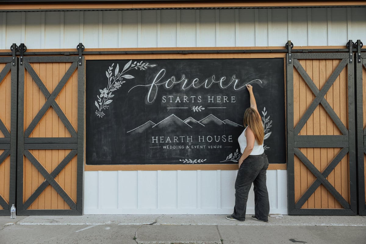 Morgan hand-lettering a large chalkboard sign on barn doors for Hearth House wedding venue, reading forever starts here with mountain illustration and botanical details