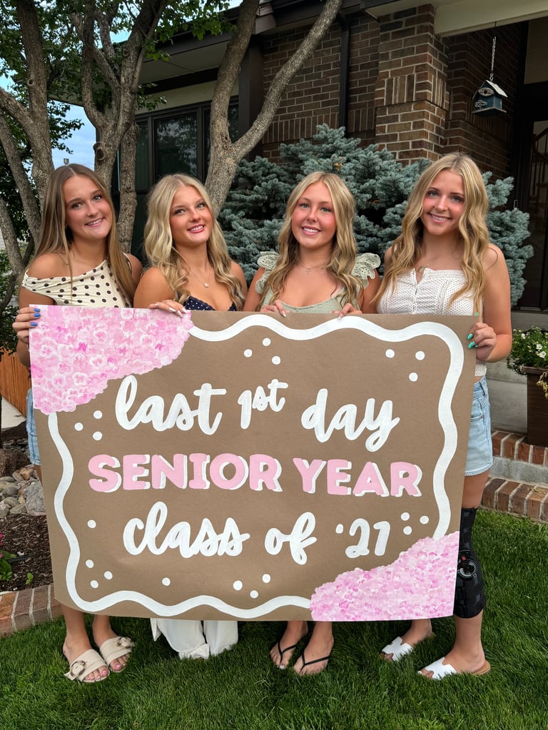 Four blonde female high school seniors holding a pink and beige sign reading last 1st day senior year class of 2027 in front of a brick house