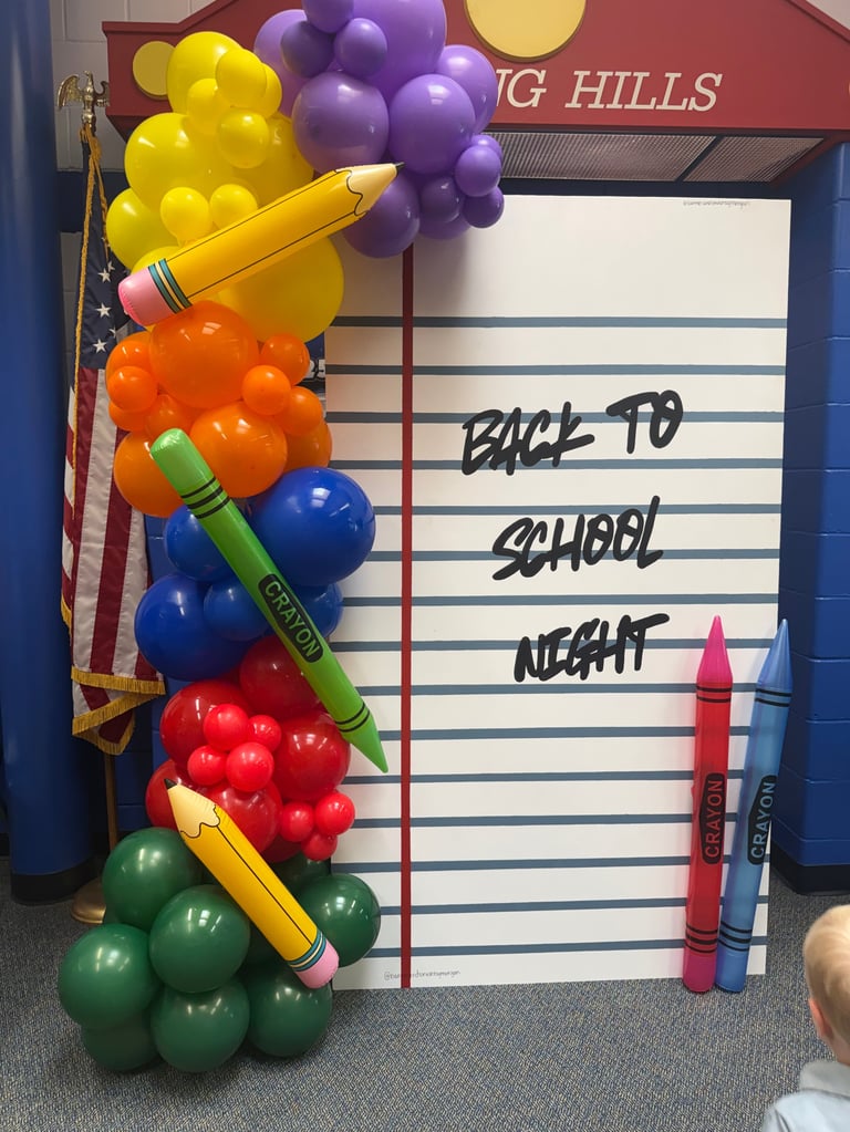 Back to school night sign decorated with colorful balloons arranged like crayons and oversized crayon props against a notepad background