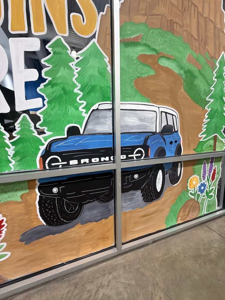 Mural of two Ford Broncos on a forest road with evergreen trees and wildflowers painted on a storefront window
