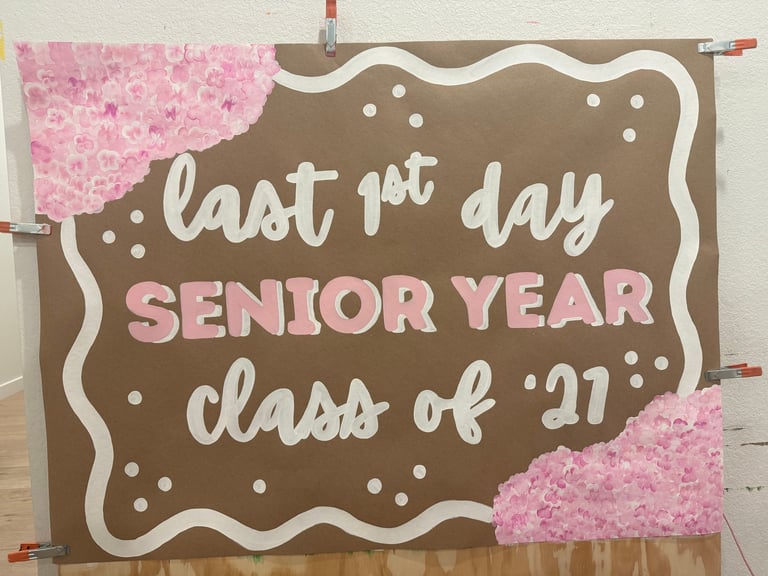 Handmade poster with brown background, white wavy border, and pink flowers reading last 1st day SENIOR YEAR class of 2027