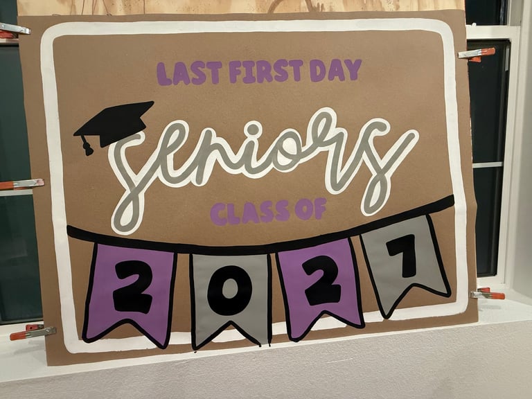 Cardboard sign reading Last First Day Seniors Class of 2027 with purple and white banner flags