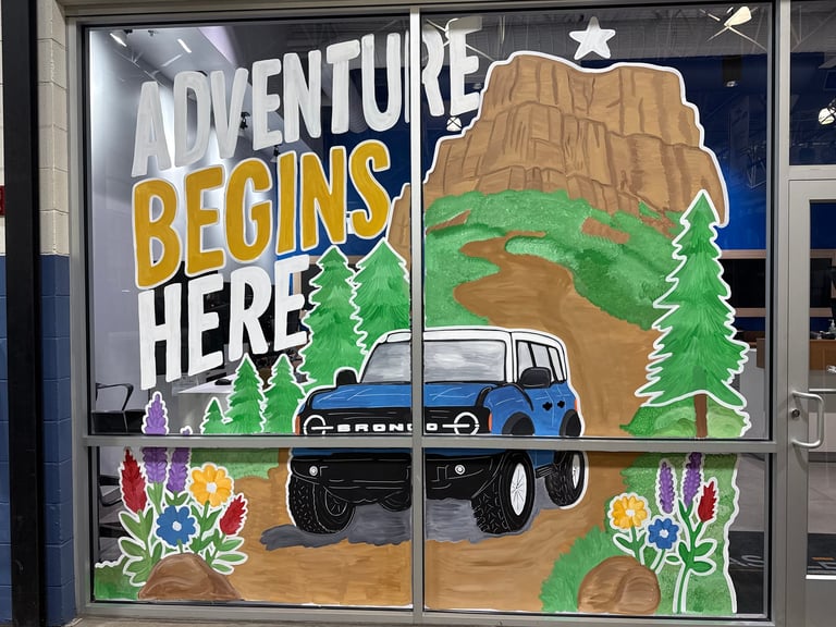 Window display with Adventure Begins Here text, illustrated blue pickup truck, desert mountains, evergreen trees, and colorful wildflowers