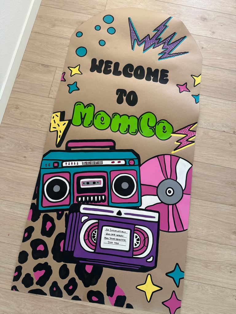 Colorful cardboard welcome sign for MomCo featuring retro boomboxes, cassette tapes, stars, lightning bolts, and leopard print designs on wood floor