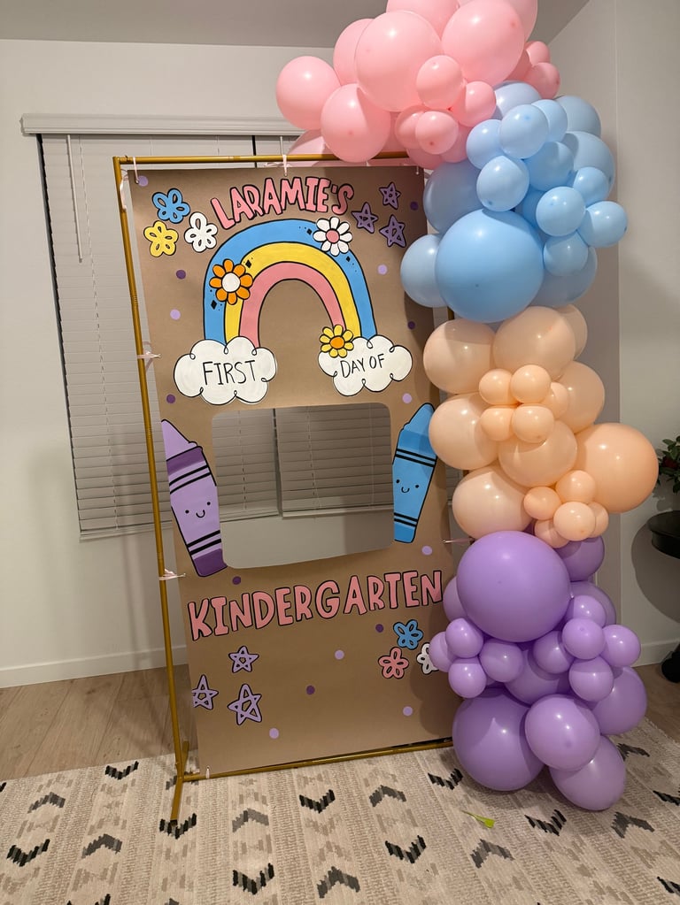Decorated cardboard photo booth frame celebrating First Day of Kindergarten with rainbow design, colorful balloons, and crayons, positioned indoors with patterned floor