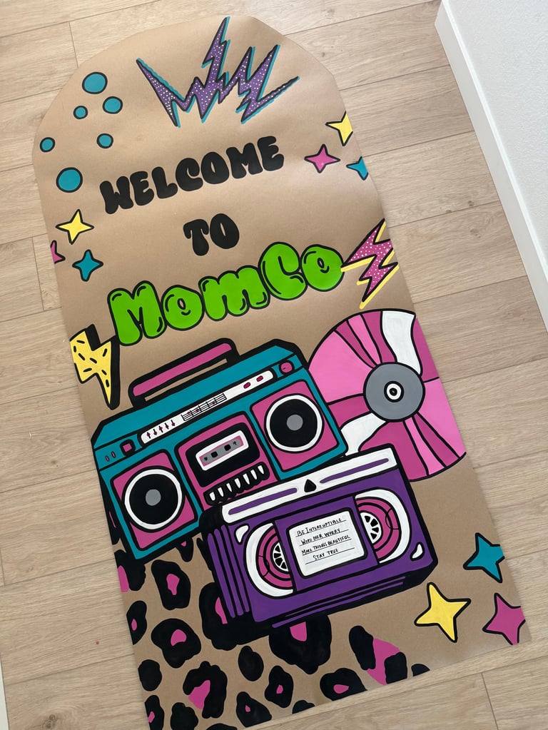 Handmade cardboard sign reading Welcome to Mom Co decorated with colorful boom boxes, lightning bolts, stars, and leopard print patterns