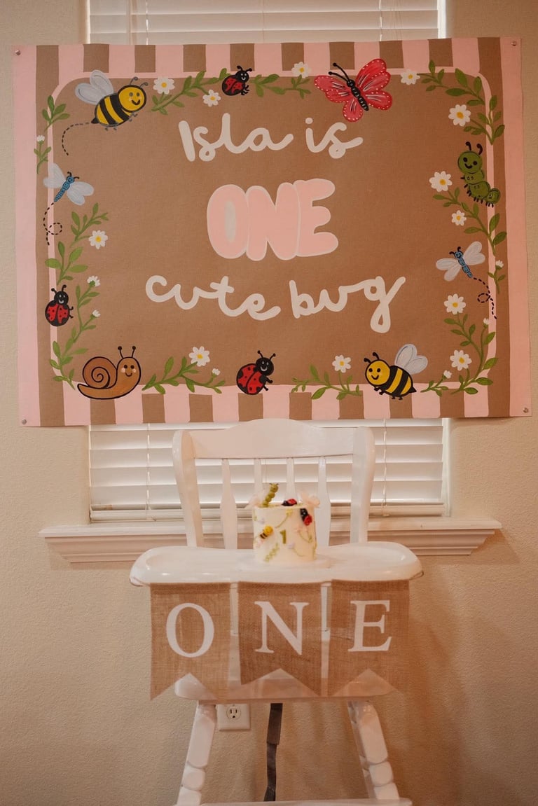 First birthday party decoration with cute bug theme featuring tan and pink signage saying Isla is ONE cute bug above a white chair decorated with ONE banner