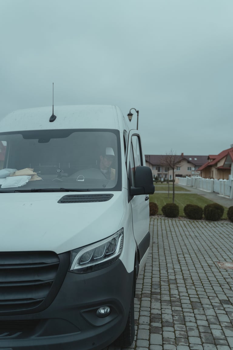 White commercial van parked on brick driveway with residential house and white fence in background under overcast sky