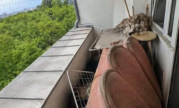 Elevated view of a residential balcony with weathered cushions, scattered debris, wire fencing, and green landscape visible below