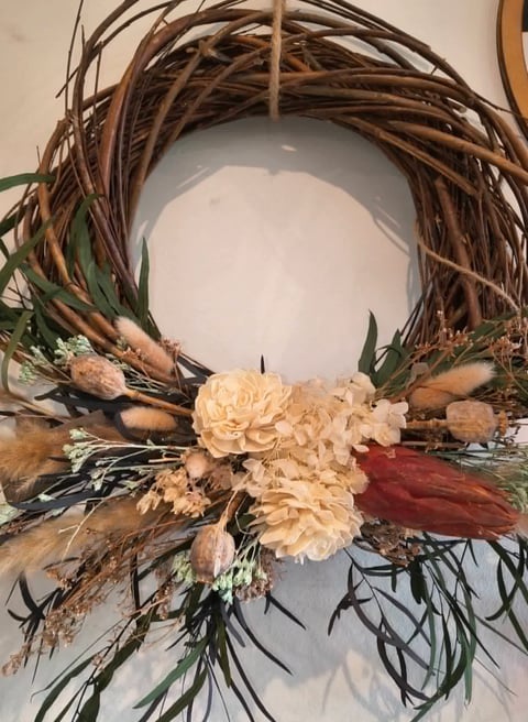 Decorative wreath made with brown woven branches, cream-colored flowers, burgundy feathers, and dried botanicals against white background