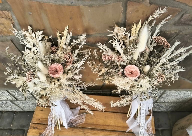 Two dried flower bouquets with pink roses, white pampas grass, and babys breath tied with white ribbons on wooden surface