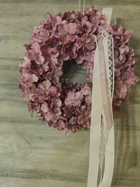Dried dusty rose hydrangea wreath with white lace ribbon hanging on wooden wall