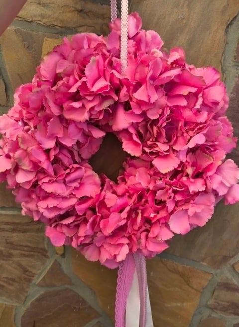 Bright pink carnation wreath with full, layered petals hanging from a pink and white ribbon bow against a wooden wall