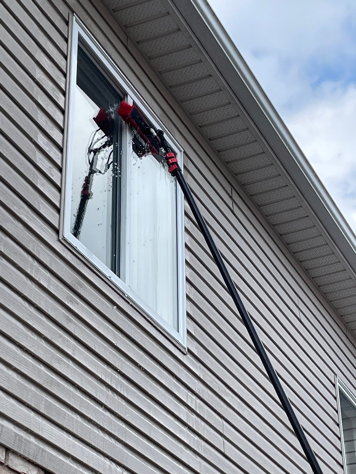 Window washer in red and black gear suspended by rope cleaning a large residential window on gray siding