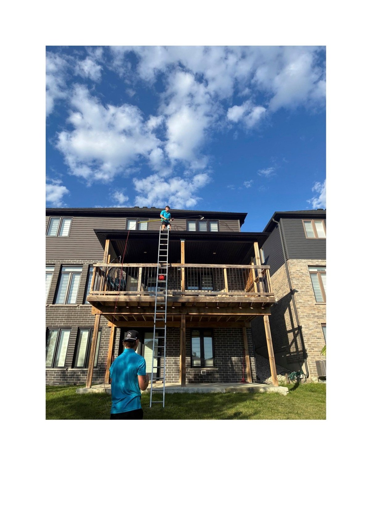Worker on ladder cleaning house exterior deck under blue sky with white clouds