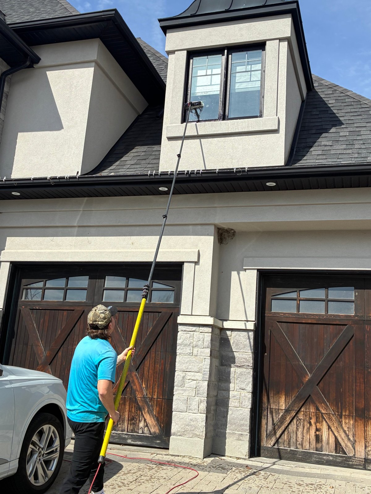 Person in blue shirt on ladder cleaning second story windows of two-story house with dark garage doors
