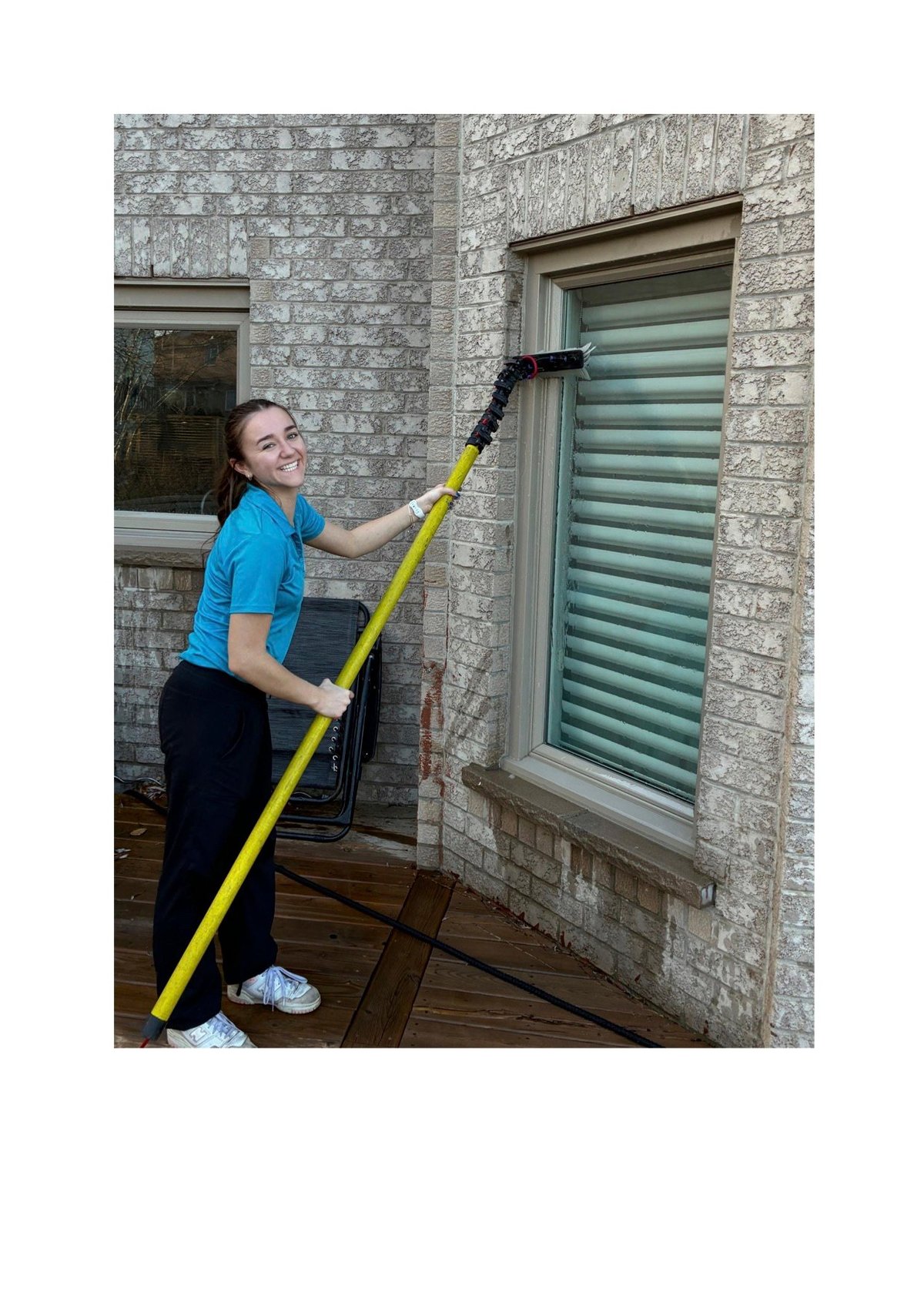 Woman in blue shirt cleaning green shuttered window with yellow extended cleaning pole on brick wall