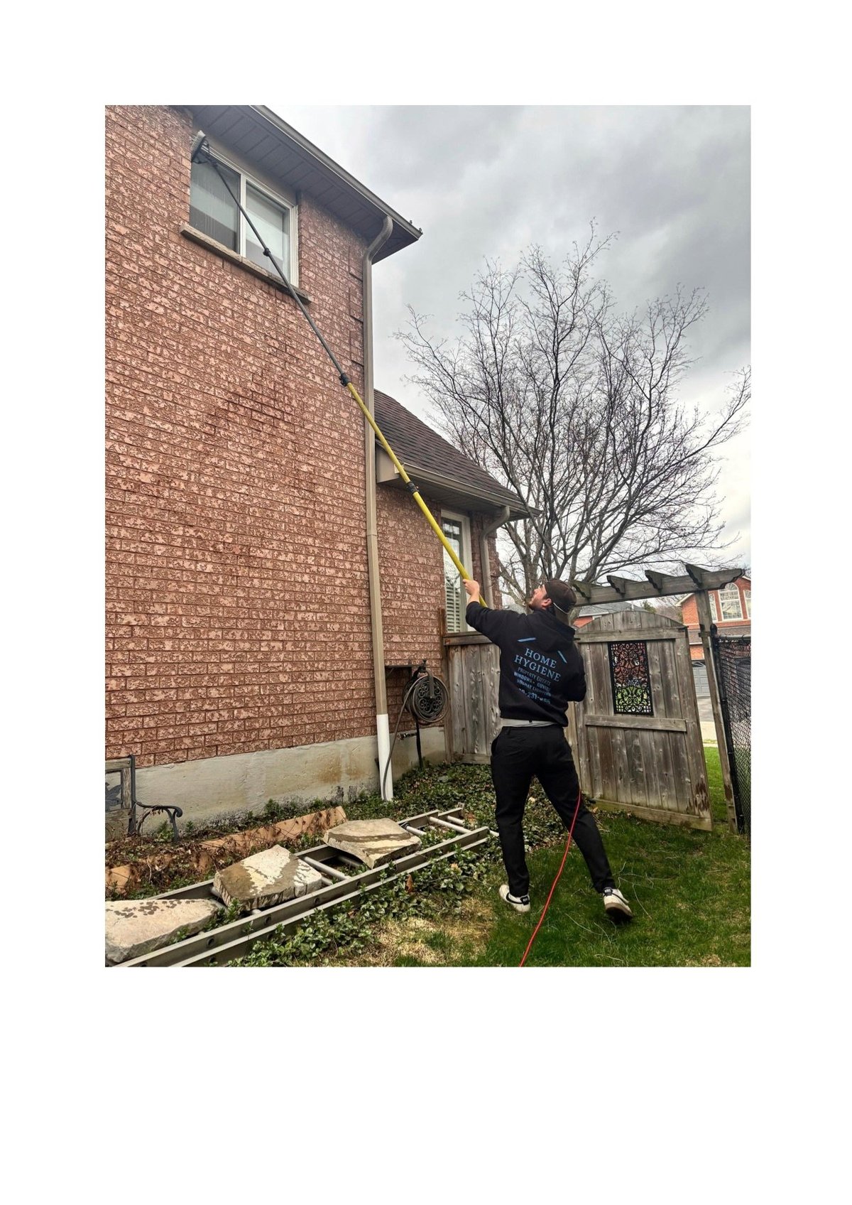 Person in black clothing standing against a brick house, reaching up toward a window with a bare tree in the background