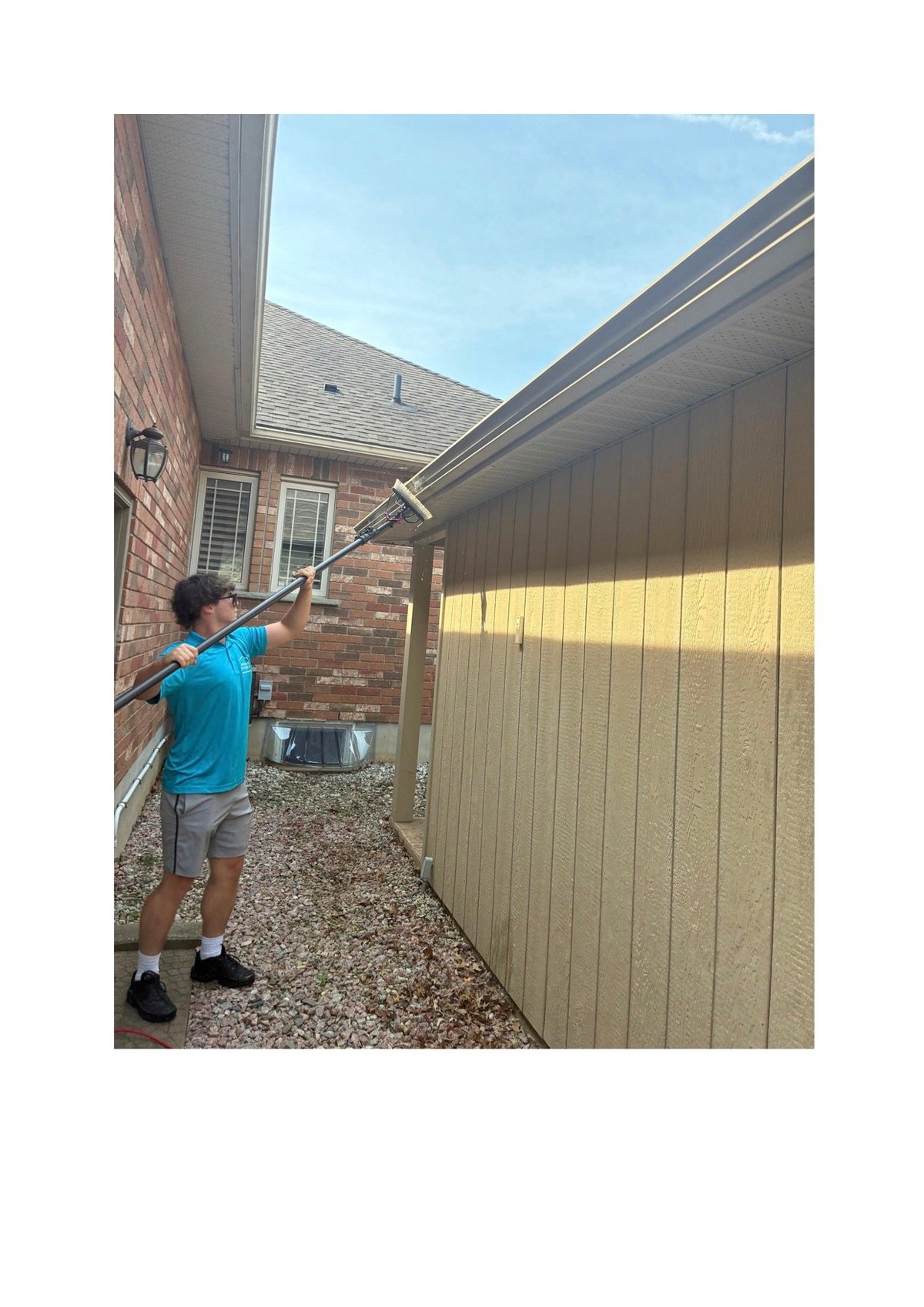 Person in blue shirt using long-handled brush to clean side of brick house with tan garage door