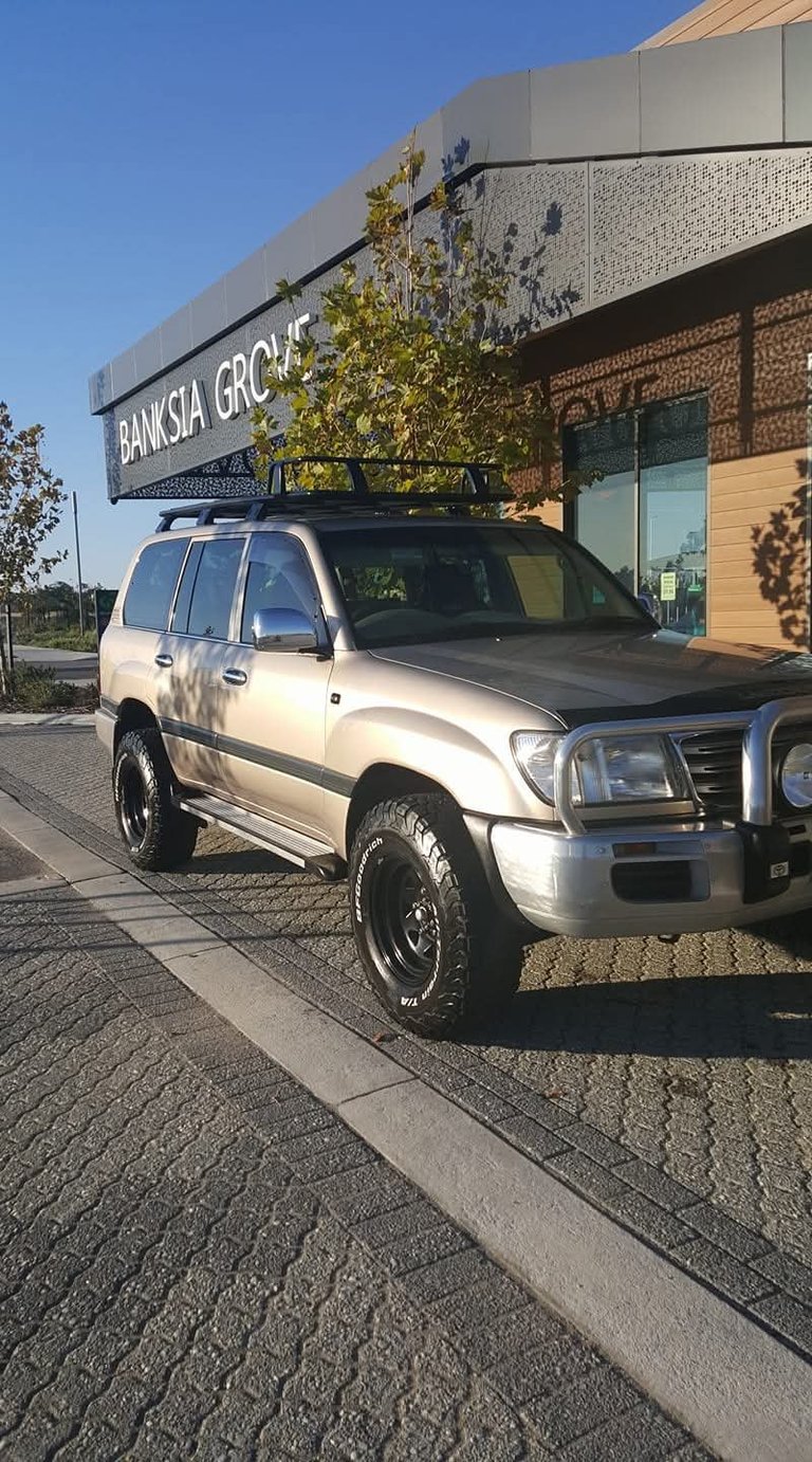 Silver Toyota Land Cruiser with black wheels parked in front of the Banksia Grocer building under a clear blue sky