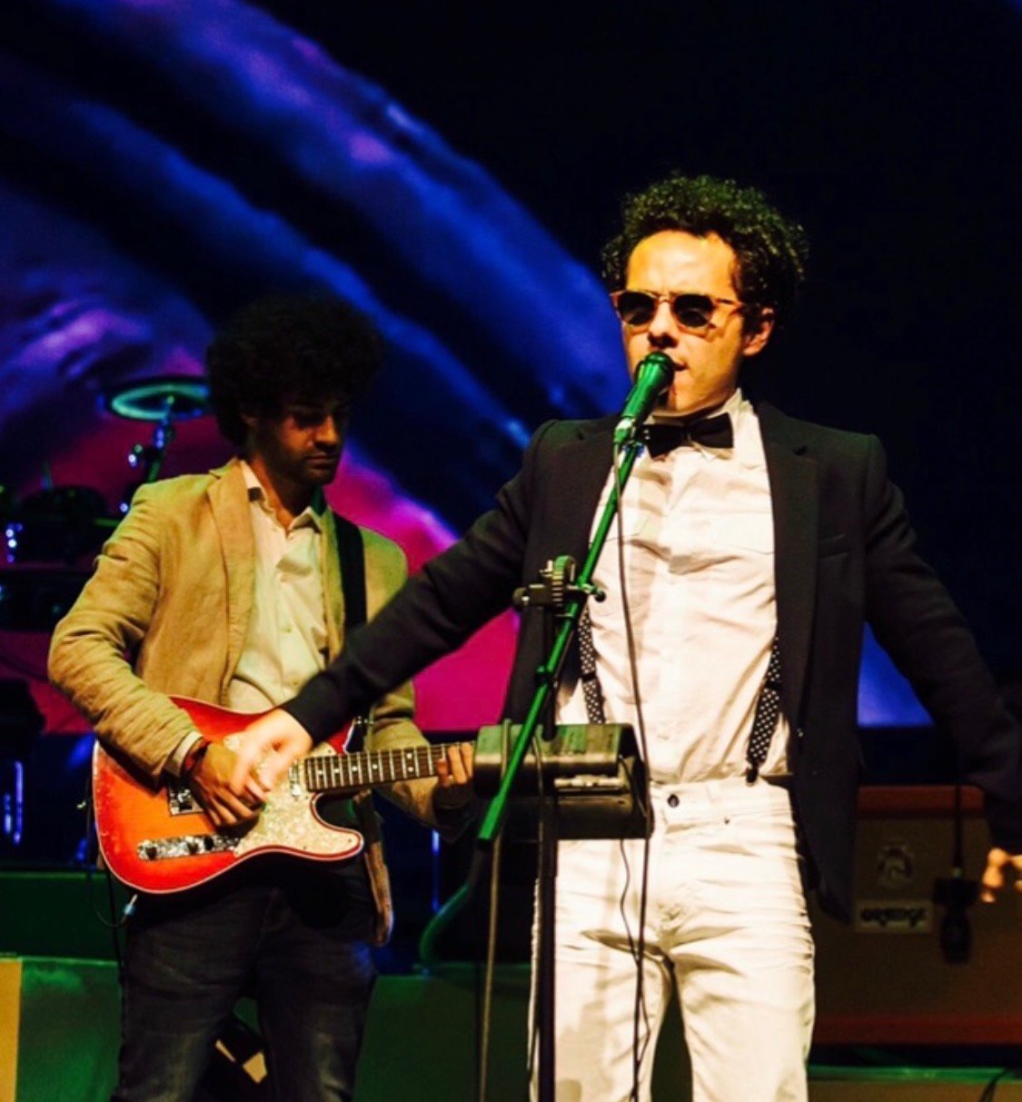 Two musicians performing on stage. Guitarist in tan jacket on left, vocalist in black blazer and sunglasses singing at microphone on right, with blue and purple stage lighting in background.