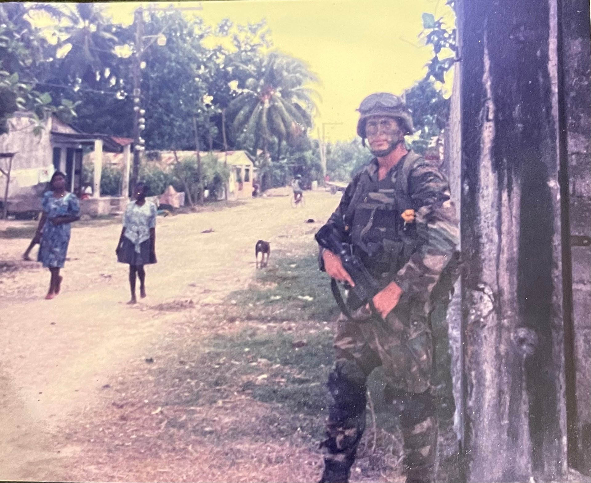Armed soldier in camouflage uniform and helmet standing against a wall on a rural village street with local civilians nearby