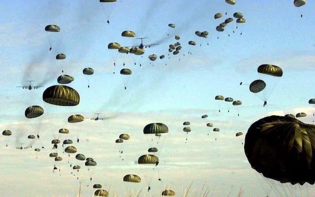 Military cargo planes with hundreds of parachutes deployed across a blue sky during a large-scale airborne operation