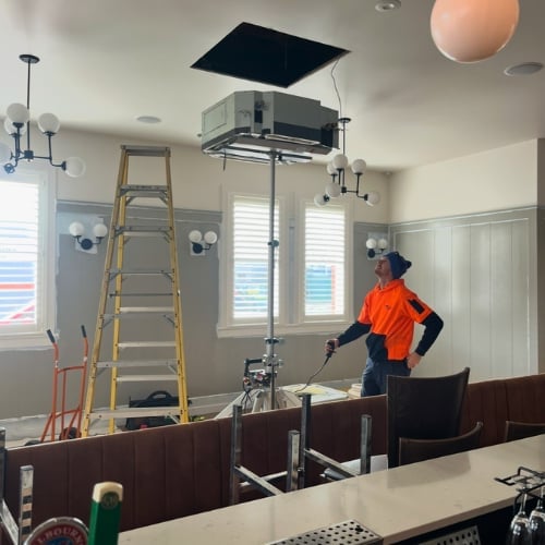 Worker in orange shirt installing ceiling-mounted HVAC unit using ladder in modern office space with white paneling