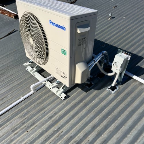 Panasonic air conditioning outdoor unit mounted on a corrugated metal roof with visible piping and cables