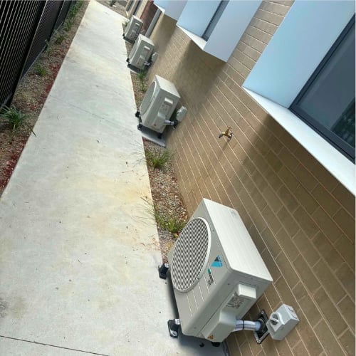 Outdoor air conditioning units mounted on a brick building wall next to a concrete sidewalk and grassy area