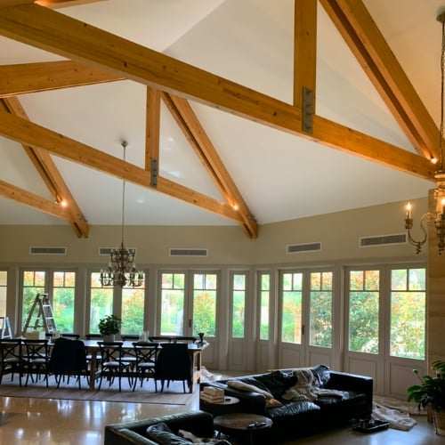 Modern open-concept living room with exposed wooden beams, vaulted ceiling, black leather furniture, and expansive wall of glass doors overlooking green landscape.