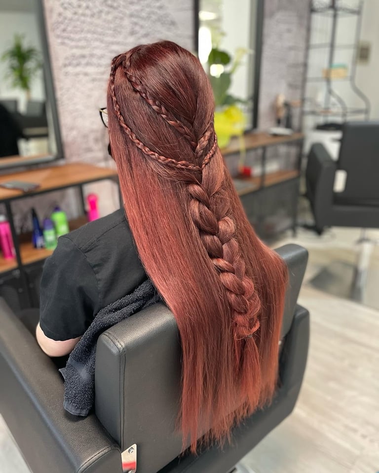 Woman with long reddish-brown hair styled with braids, seated in a salon chair viewed from behind