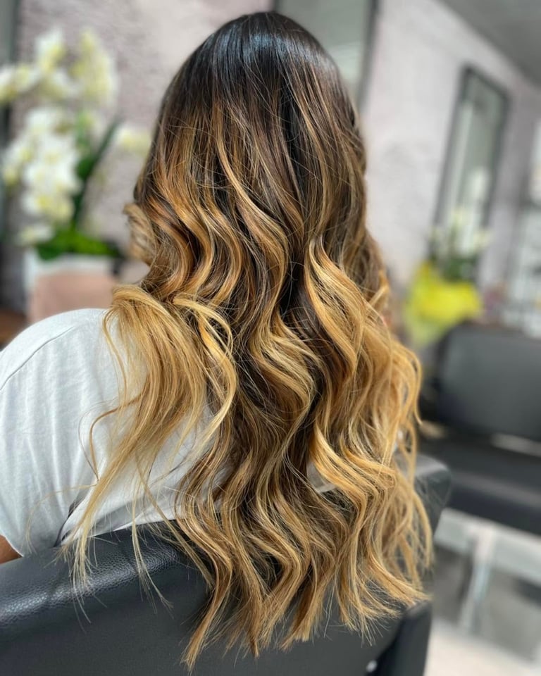 Woman with long wavy hair featuring dark roots and golden blonde balayage highlights, viewed from behind