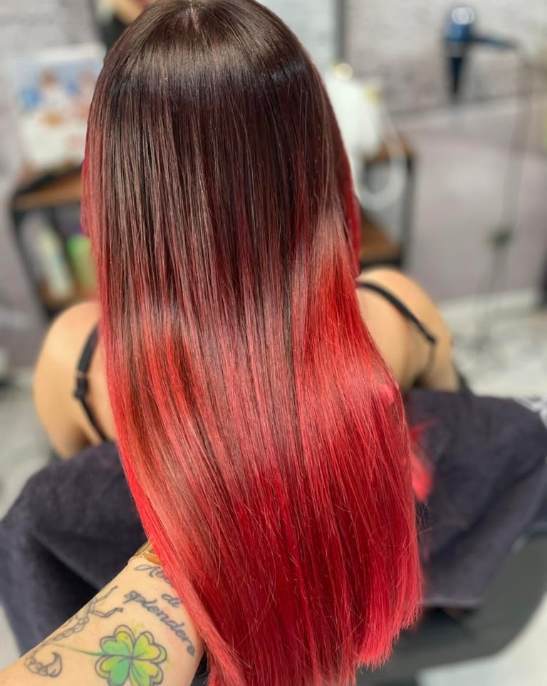 Back view of person with long straight hair featuring ombré color gradient from brown to vibrant red, wearing black sleeveless top with visible shoulder tattoo in a salon setting