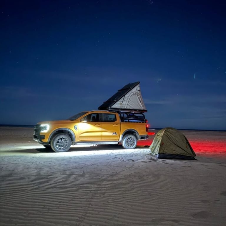 Gold pickup truck on beach at night with roof tent open and small tent nearby, illuminated by headlights and red accent lighting