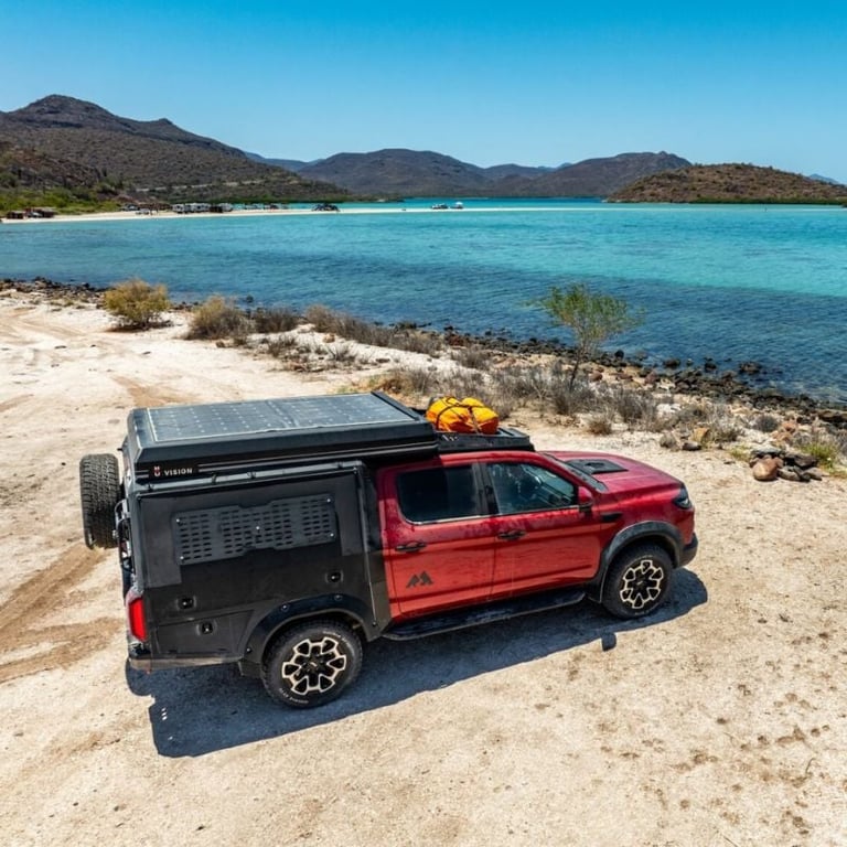 Red pickup truck with rooftop cargo carrier and solar panel parked on sandy beach with turquoise water and mountains in background