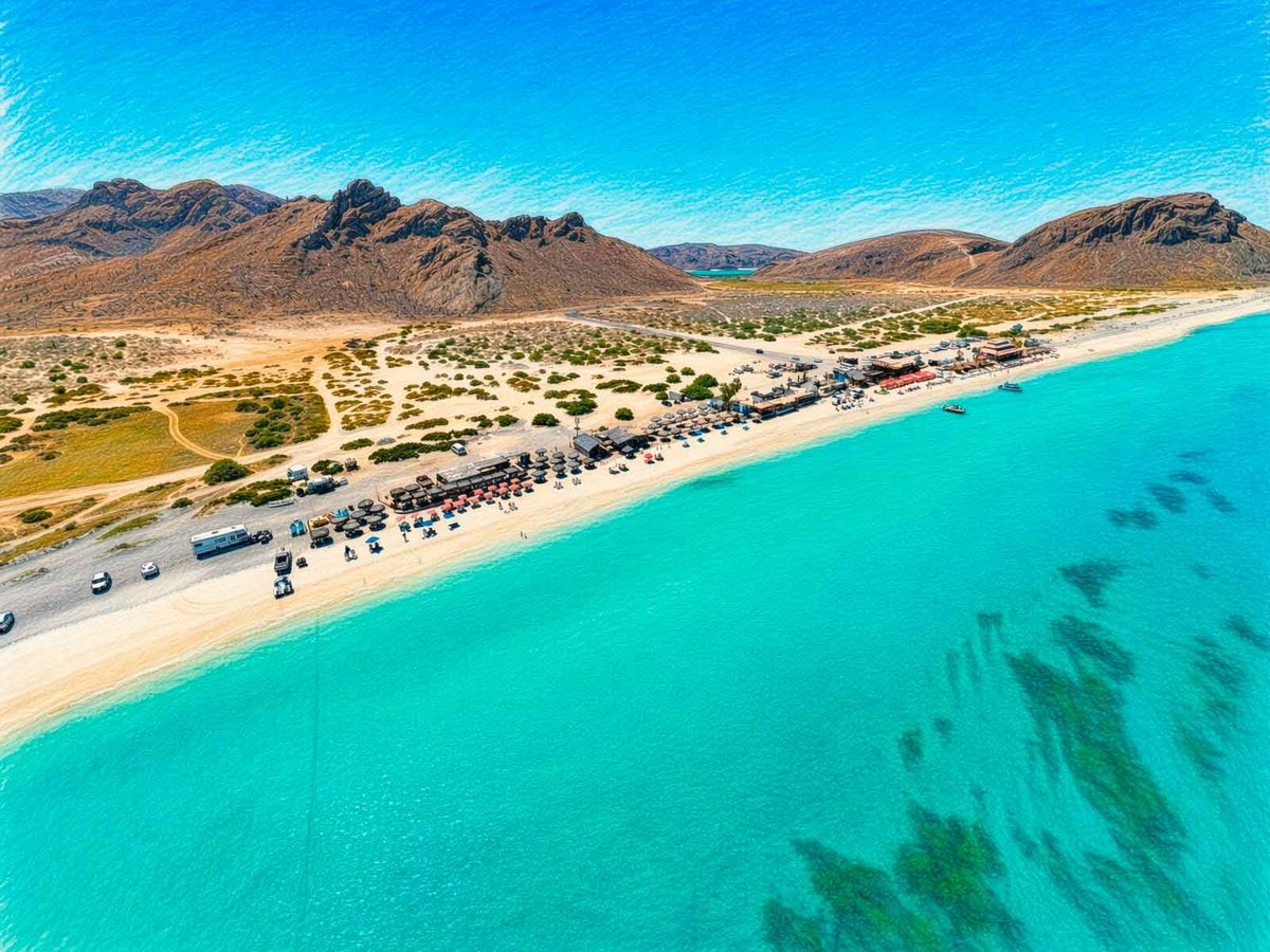 Vista aérea de Playa El Tecolote: extensa playa de arena dorada con autos estacionados, palapas y aguas turquesas del Mar de Cortés, flanqueada por montañas desérticas