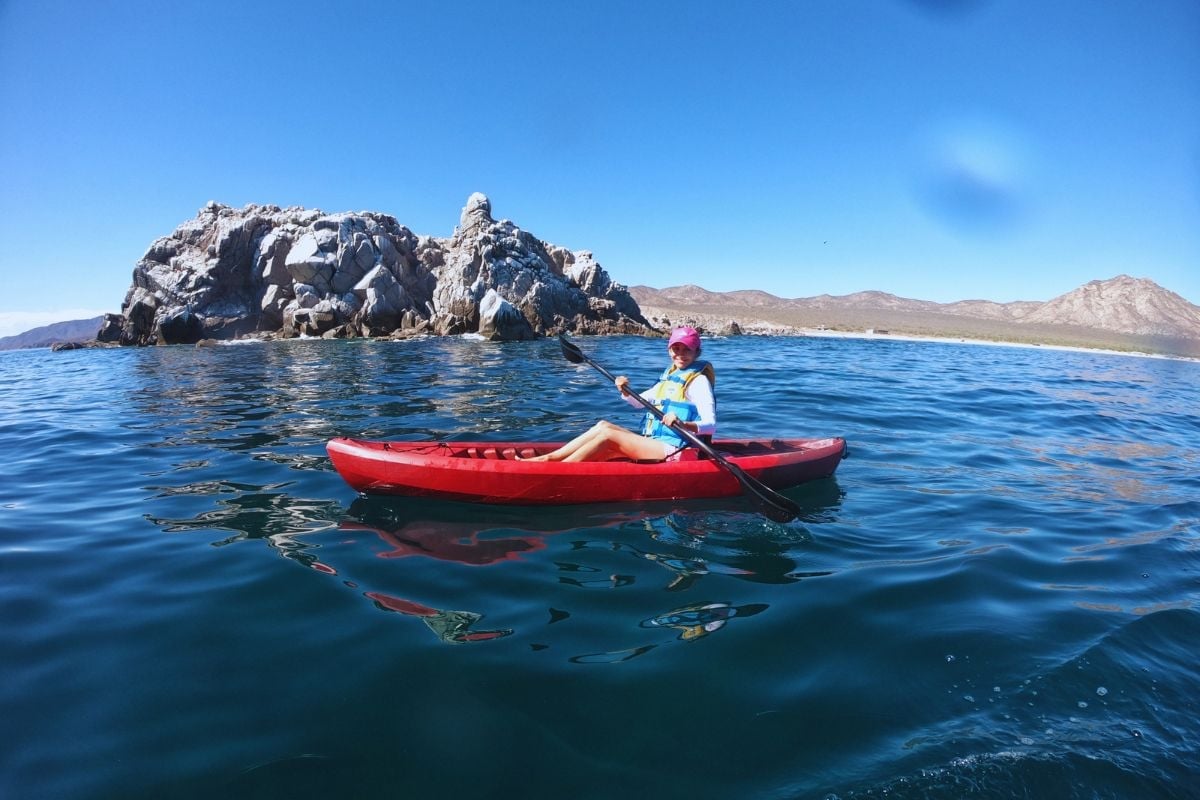 Person kayaking in a red kayak on blue water with rocky island and mountains in the background