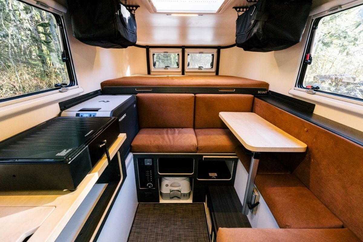 Interior of a vintage travel trailer with wood paneling, orange seating, compact kitchenette, and windows showing forest scenery outside