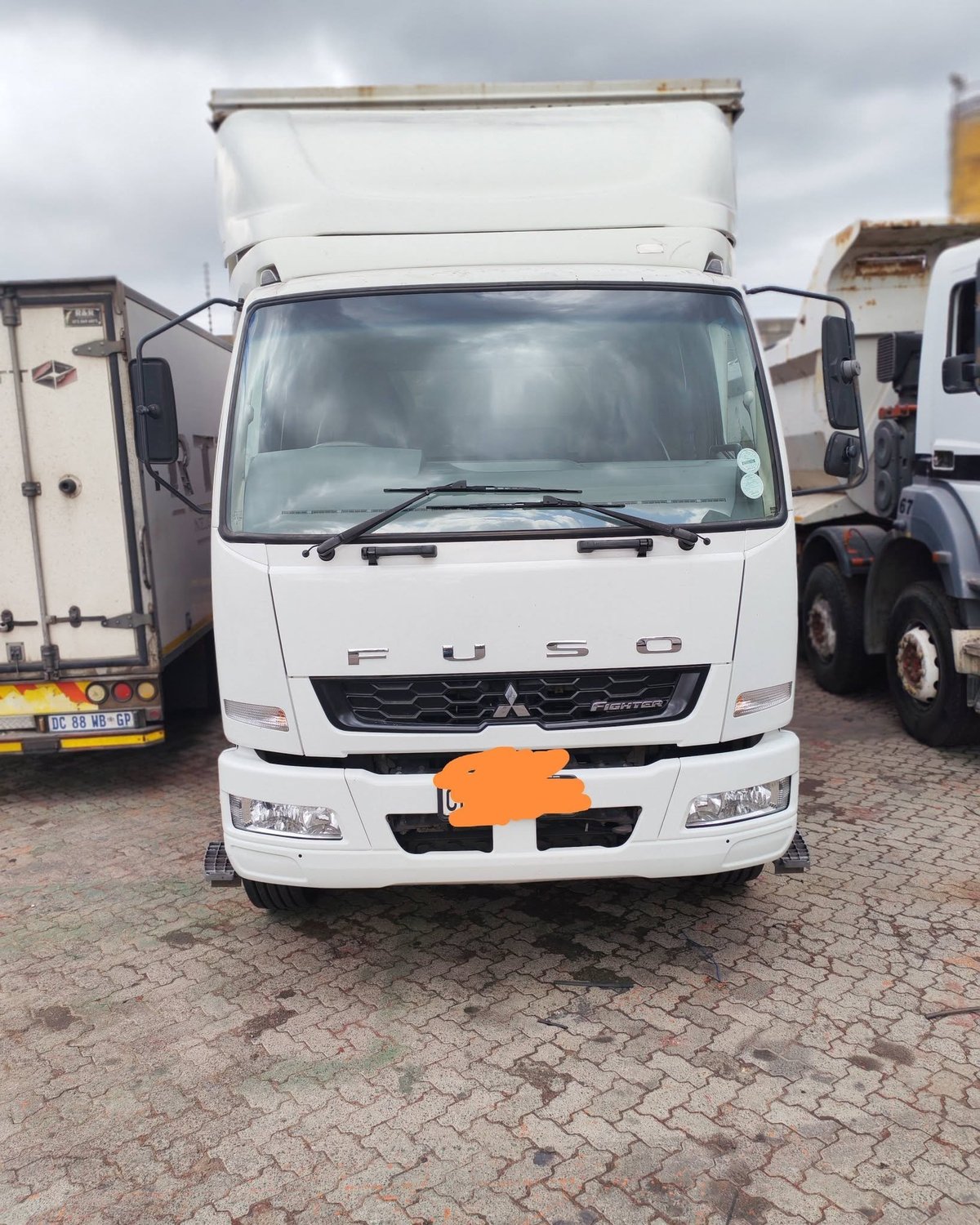 White Fuso commercial truck with orange badge parked in lot with other vehicles and cloudy sky