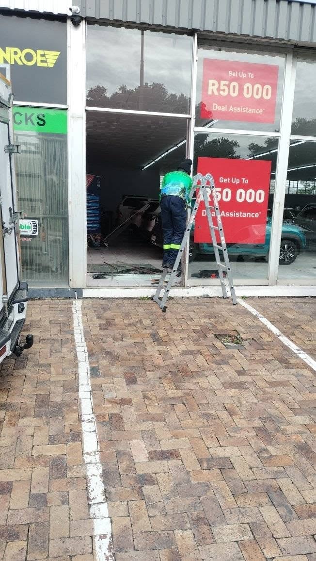 247 Affordable Glass Repairs worker in a green safety shirt standing on an aluminium step ladder, fitting red R50 000 Deal Assistance promotional banners onto a freshly installed auto repair shop shopfront window.
