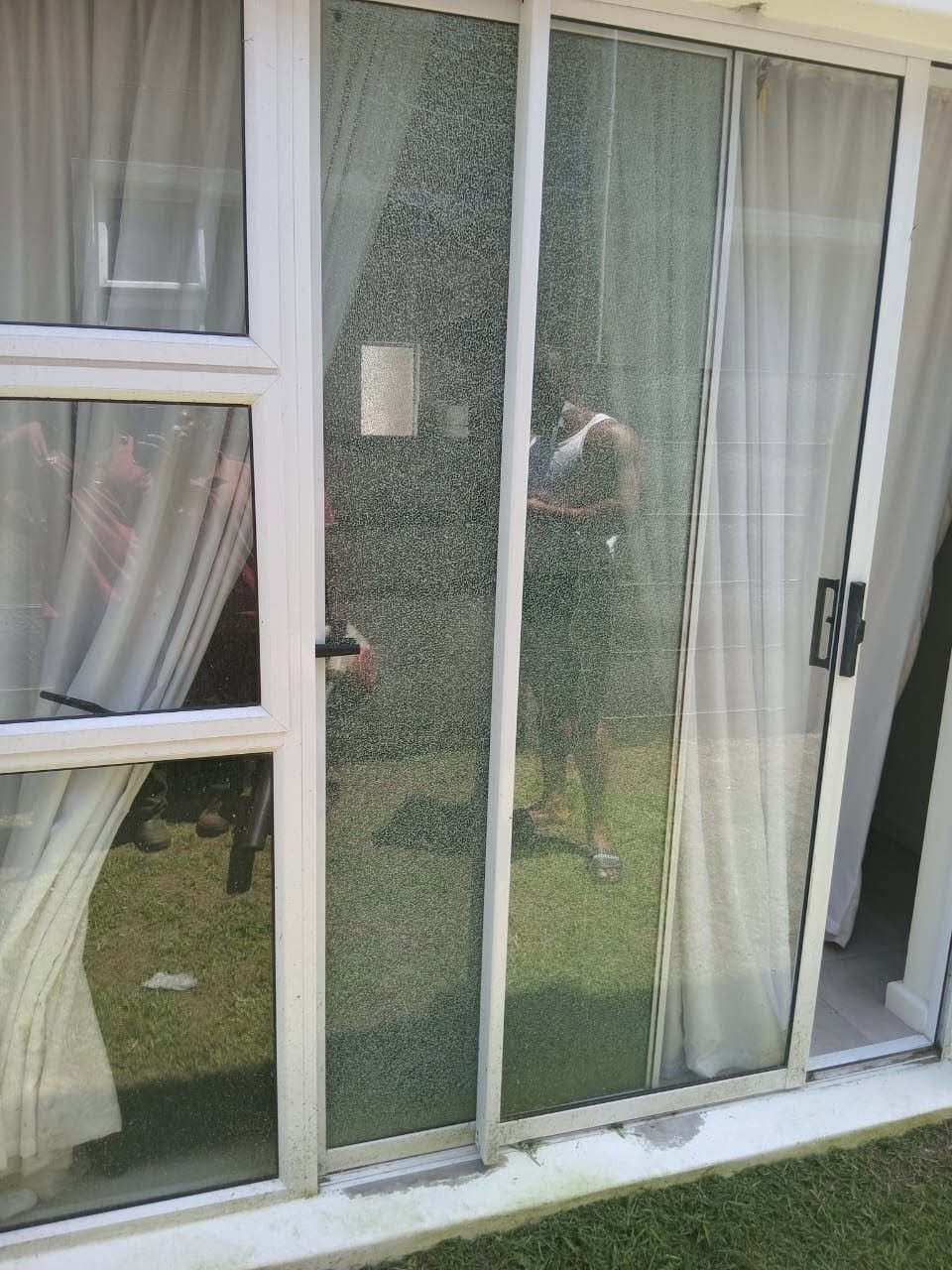 Sliding glass patio door with white aluminium frame, shattered glass and a technician in a high-vis vest visible through the broken pane, with the lawn and garden seen outside.