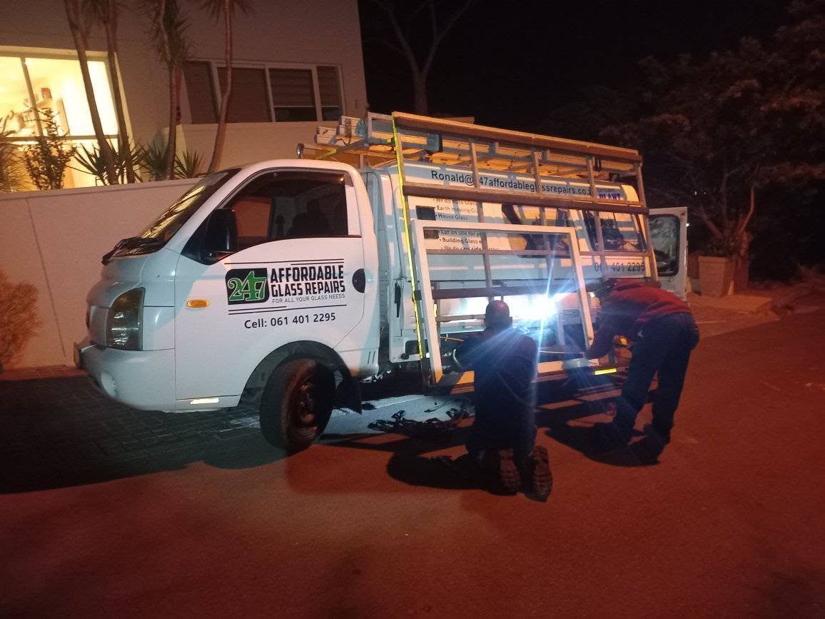 247 Affordable Glass Repairs mobile workshop van parked outside a Devil's Peak home at night with two technicians loading glass and equipment from the open cargo bay, the company branding (247 Affordable Glass Repairs, For All Your Glass Needs, Cell 061 401 2295) and email Ronald@247affordableglassrepairs.co.za clearly visible on the door.