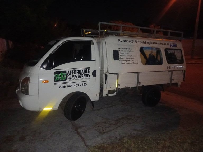 247 Affordable Glass Repairs mobile workshop van parked on a Devil's Peak residential street at night, with the yellow puddle light under the door on, the company branding (247 Affordable Glass Repairs — For All Your Glass Needs — Cell 061 401 2295) clearly visible on the door, and a list of services (Auto Glass, Earth moving Glass, House Glass, Service Sliding Doors, Cut on site for you, Building Glass, side mirrors) printed on the cargo box alongside the email Ronald@247affordableglassrepairs.co.za.