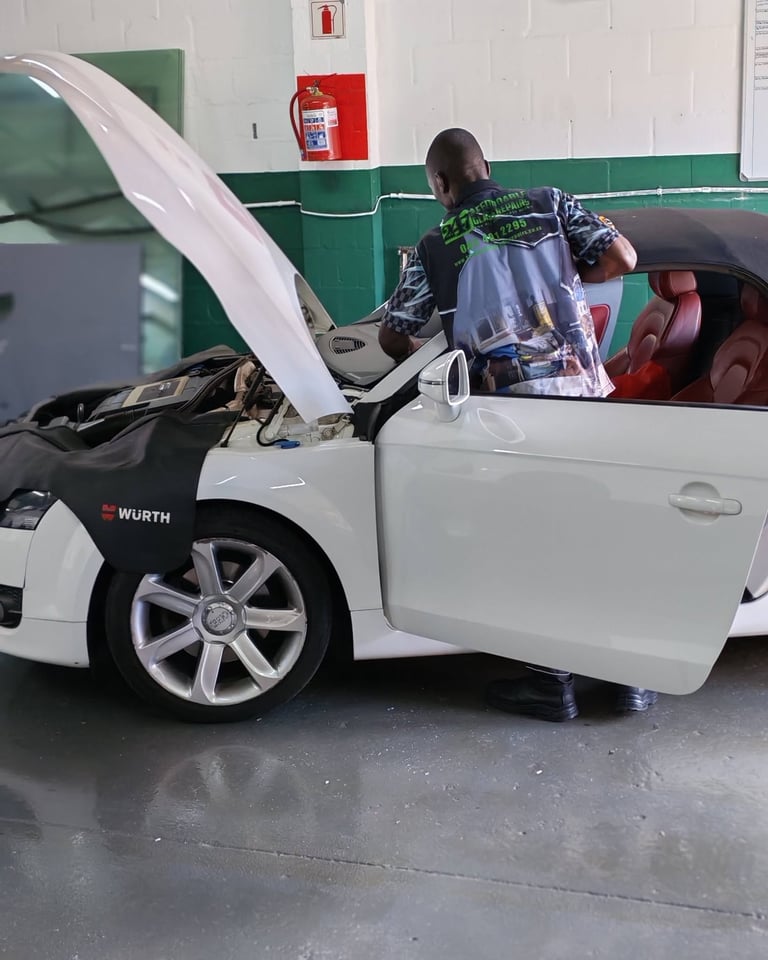 247 Affordable Glass Repairs mechanic in branded shirt working on a white Audi TT with the bonnet open and red leather interior visible inside the green-and-white garage workshop.