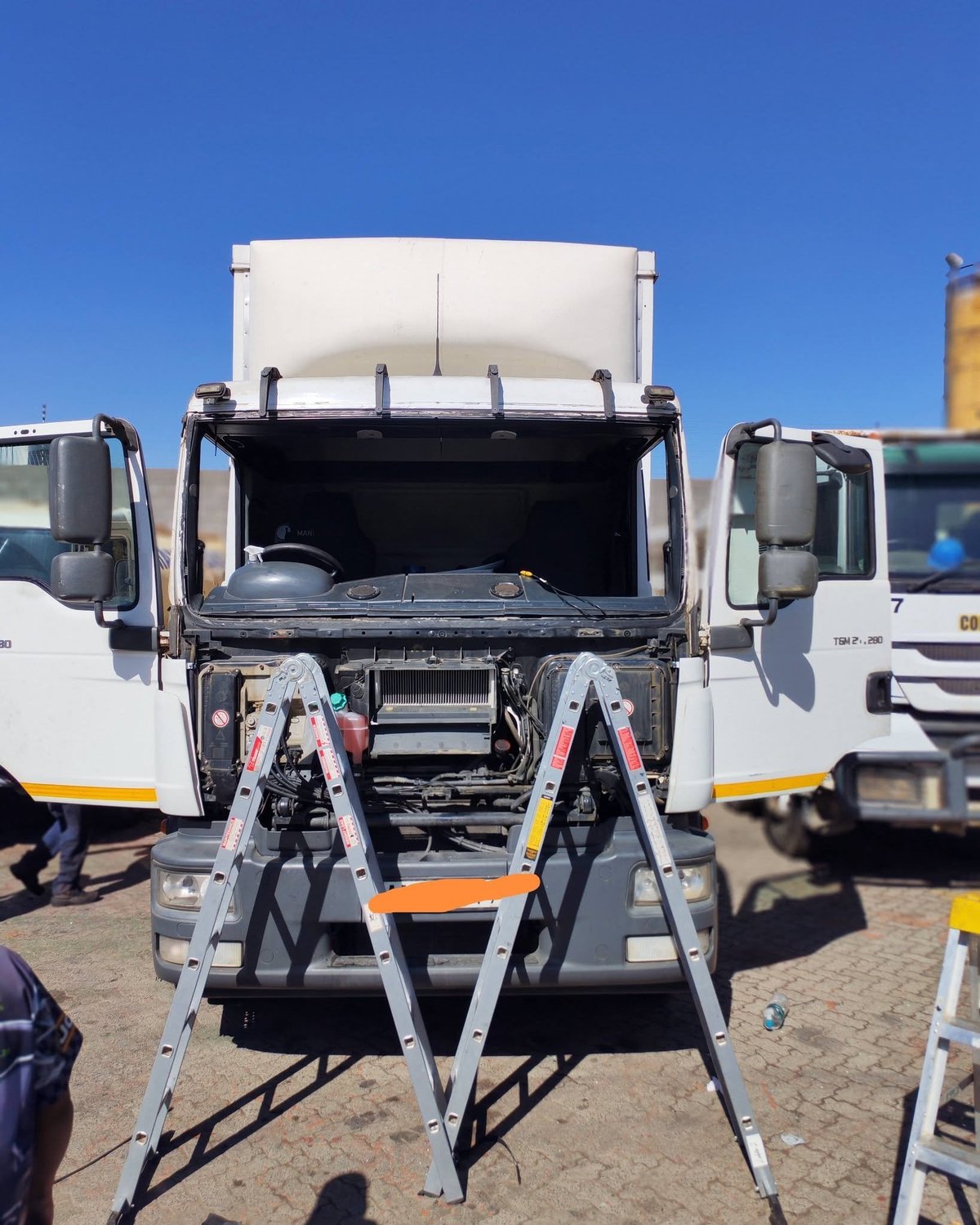 Open cargo truck with hood raised and aluminum ladder leaning against engine, in outdoor parking lot