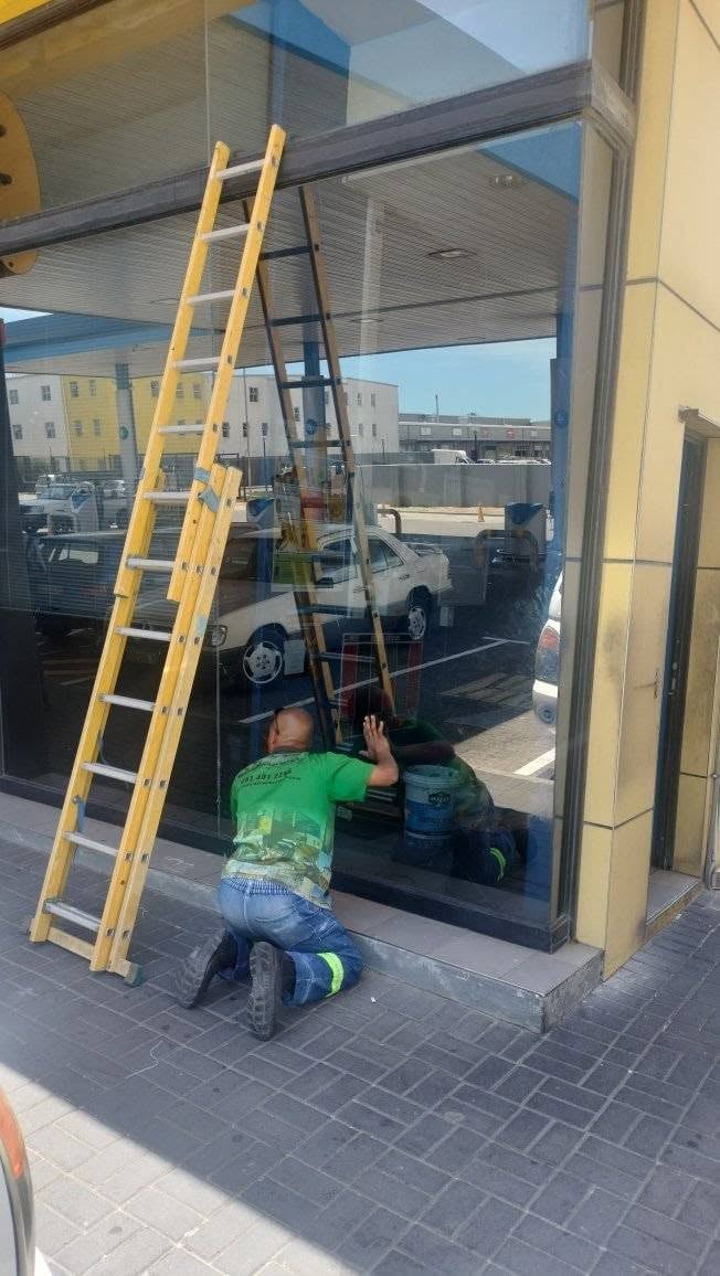Worker in green shirt fitting a large glass storefront window with a yellow ladder leaning against the frame